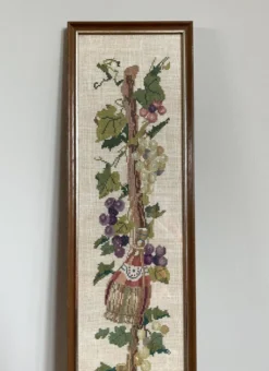 Broderie néerlandaise encadrée représentant une vigne avec des raisins, des bouteilles de vin et des paniers.