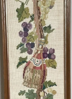 Broderie néerlandaise encadrée représentant une vigne avec des raisins, des bouteilles de vin et des paniers.