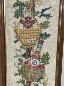 Broderie néerlandaise encadrée représentant une vigne avec des raisins, des bouteilles de vin et des paniers.