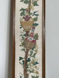 Broderie néerlandaise encadrée représentant une vigne avec des raisins, des bouteilles de vin et des paniers.