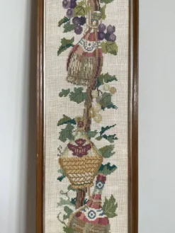 Broderie néerlandaise encadrée représentant une vigne avec des raisins, des bouteilles de vin et des paniers.
